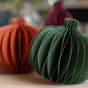 May include: Three spherical paper decorations shaped like pumpkins, featuring a ribbed, accordion-style construction. The foreground displays a dark green pumpkin, while an orange pumpkin and a deep burgundy pumpkin are positioned behind it.