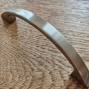May include: A silver-toned, curved cabinet pull with a rectangular base. The pull is shown on a wood surface.