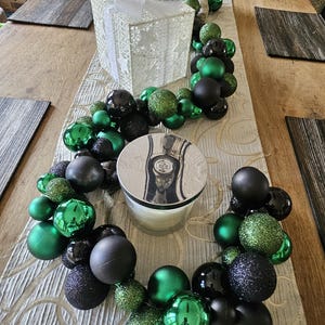 May include: A Christmas centerpiece featuring a white gift box with a pinecone and greenery, surrounded by a garland of green and black ornaments. A candle sits in the center of the garland on a textured runner.