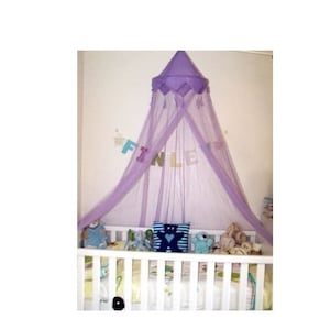 May include: A purple canopy mosquito net with tassels hangs over a white cot with stuffed toys and a blue pillow with a white bear design. The cot is in a room with white walls and a banner that says "FINLEY" on it.