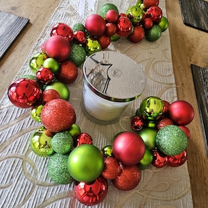 May include: A festive Christmas table centerpiece featuring a white candle with a silver lid, surrounded by a garland of red, green, and glittery ornaments. The arrangement sits on a white lace table runner.