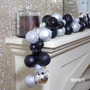 May include: A decorative garland of silver, black, and glittery ornaments draped across a white mantel. A glass jar candle sits on the left. The garland is a mix of matte and shiny spheres, creating a festive holiday display.