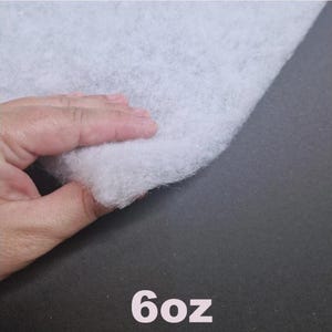 May include: A close-up of a white, fluffy fabric, possibly batting or wadding, is held by a hand. The text "6oz" is printed in white at the bottom of the image, indicating the weight of the material.