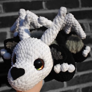 Puede incluir: Una cabeza de ciervo de ganchillo hecha a mano con cuernos blancos, una cara blanca y detalles en negro. El ciervo tiene ojos grandes y expresivos con un degradado de colores. La nariz del ciervo es en forma de corazón negro. El fondo es una pared de ladrillos.