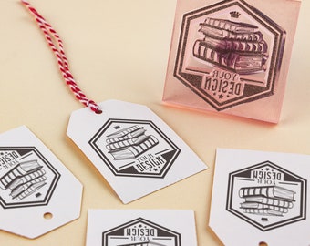May include: Four white gift tags with a black and white illustration of a stack of books with the text "Your Design" in a hexagon shape. A pink rubber stamp with the same design is also shown.