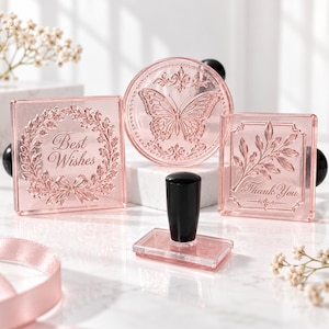 May include: Three translucent pink acrylic stamps with black handles. One square stamp features a wreath and the text Best Wishes. One circular stamp displays a butterfly. One square stamp shows a leafy branch and the text Thank You.