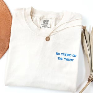 May include: Cream-colored t-shirt with the text "NO CRYING ON THE YACHT" in blue. A gold-colored necklace with a round pendant is draped across the shirt. The shirt has a Comfort Colors tag.