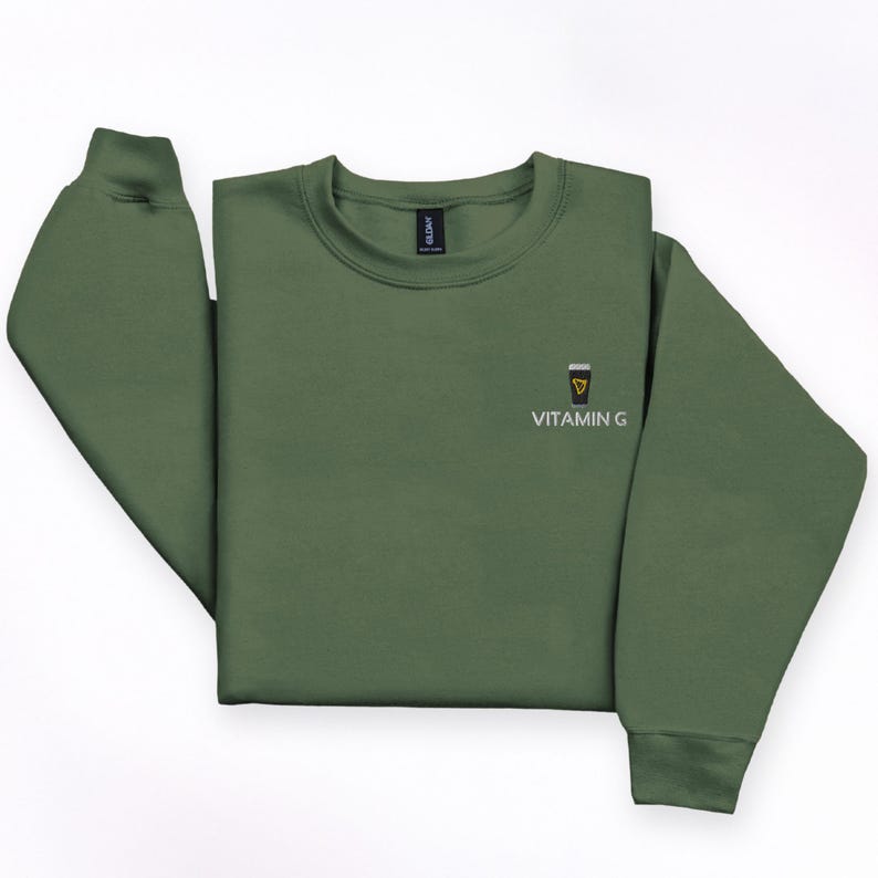 May include: Olive green sweatshirt with a crew neck and long sleeves. The front features a small graphic of a pint of beer with a yellow triangle and the text "VITAMIN G" in white.