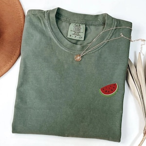May include: Olive green Comfort Colors t-shirt with a small embroidered watermelon slice on the front. A delicate silver necklace with a pendant rests on the shirt. A brown hat is visible in the upper left corner.
