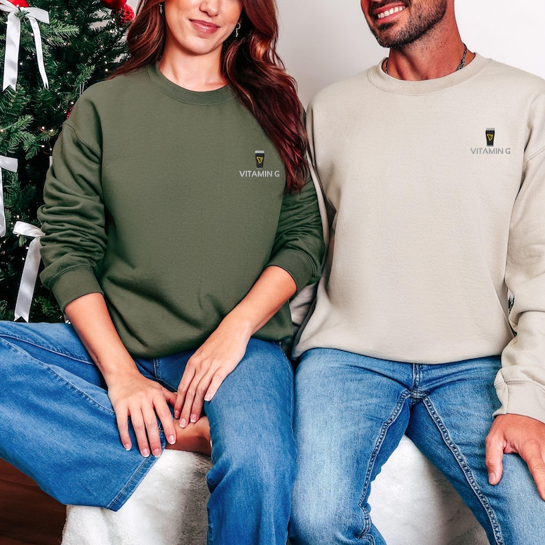 May include: Two people wearing matching sweatshirts. One is olive green, the other is beige. Both sweatshirts have the text "VITAMIN G" and a small graphic on the chest. They are wearing blue jeans.