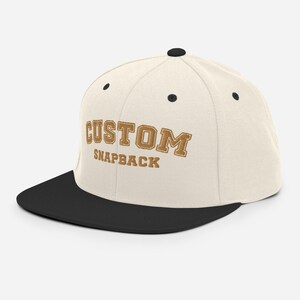 Pode incluir: Um boné snapback personalizado bege e preto com o texto "CUSTOM SNAPBACK" bordado em marrom na frente.