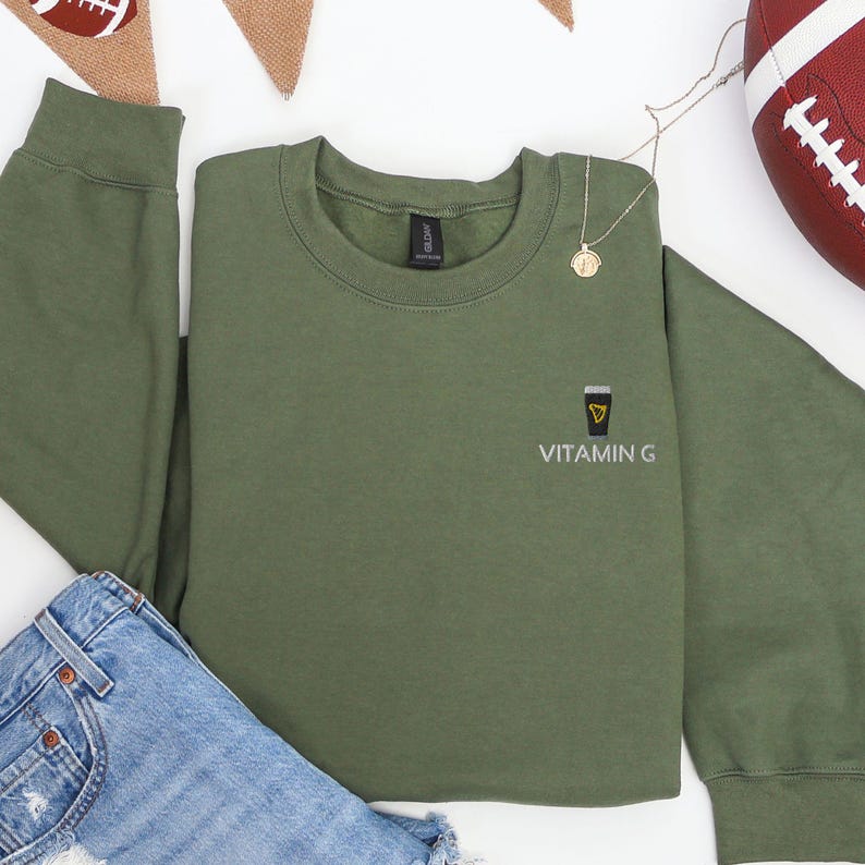 May include: An olive green sweatshirt with the text "VITAMIN G" and a small graphic of a coffee cup. A gold necklace with a pendant is on the sweatshirt. A pair of blue jeans and a football are also in the image.