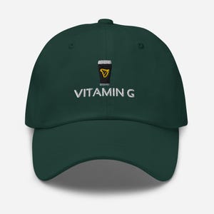 May include: A dark green baseball cap with a curved brim. The cap features an embroidered design of a black and gold pint of stout and the words "VITAMIN G" in white lettering. The cap is a casual accessory.