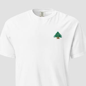 May include: White t-shirt with a small embroidered green and brown tree design on the left chest. The shirt has a crew neck and short sleeves. The fabric appears to be a soft, comfortable material.