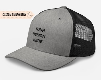 May include: A gray and black trucker hat with a black mesh back. The front of the hat has the text "YOUR DESIGN HERE" embroidered in black.
