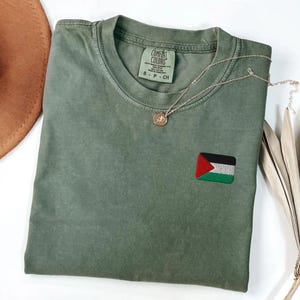 May include: An olive green Comfort Colors t-shirt with a small embroidered Palestinian flag patch. A delicate silver necklace with a pendant rests on the shirt. The shirt is folded, with a brown hat visible in the background.