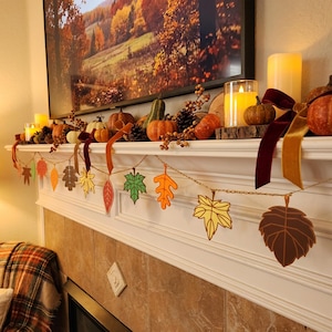 May include: A decorative garland featuring various paper cutouts of autumn leaves in shades of orange, yellow, and brown, strung along a thin cord. The garland is displayed on a white fireplace mantel adorned with pumpkins, candles, and ribbons.