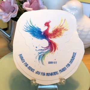 May include: A white sticker featuring a colorful phoenix design with blue, red, and yellow hues. The sticker has the text "BEAUTY FOR ASHES. JOY FOR MOURNING. PRAISE FOR HEAVINESS." and "ISAIAH 61:3". The sticker is displayed on a clear stand.