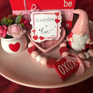 May include: A pink and red Valentine's Day themed table setting with a gnome, a heart-shaped dish, a wooden sign that says "be my one", a wooden sign that says "XOXO", and a sticker that says "Do everything in love 1 Cor 16:14".