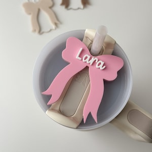 Könnte beinhalten: Rosa Schleifenförmiges Namensschild mit dem Namen "Lara" in weißen Buchstaben. Das Schild ist an einem weißen Deckel mit einem Strohhalm befestigt.