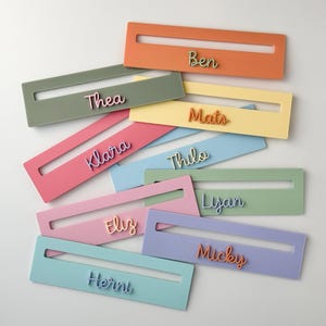 May include: A selection of pastel-coloured name tags, including pink, green, and blue. Each tag has a name in a contrasting colour, such as orange or purple. The rectangular tags have a cutout. Names include "Thea", "Klara", "Eliz", "Henni", "Ben", "Mats", "Thilo", "Lyan", and "Micky".