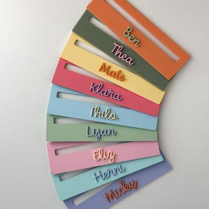 May include: A collection of colourful name tags in shades of orange, green, yellow, pink, and blue. Each tag displays a different name in a matching colour. The tags are arranged in a semi-circular pattern against a white background.