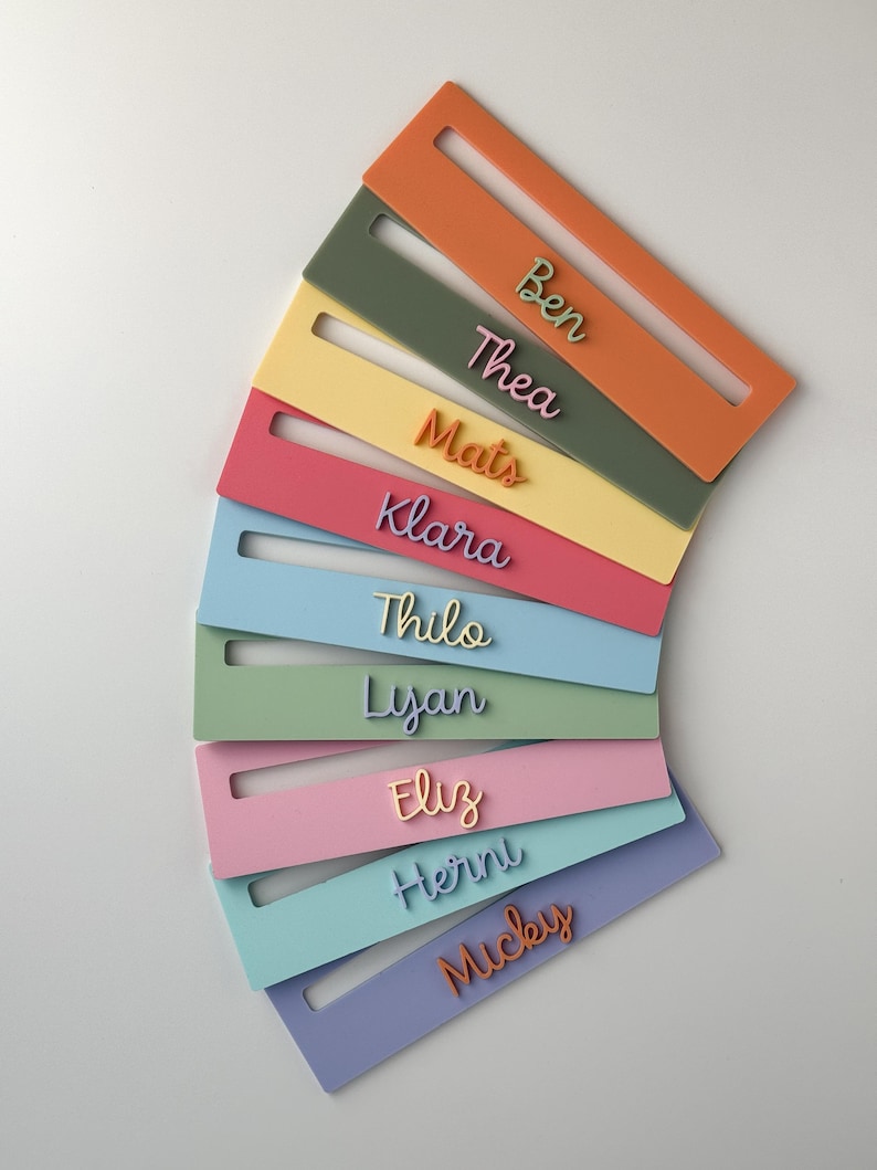 May include: A collection of colourful name tags in shades of orange, green, yellow, pink, and blue. Each tag displays a different name in a matching colour. The tags are arranged in a semi-circular pattern against a white background.