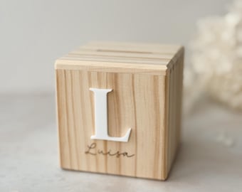 May include: Wooden piggy bank with a white letter L and the name Luisa engraved on the front. The piggy bank is sitting on a light gray surface with two gold coins beside it.