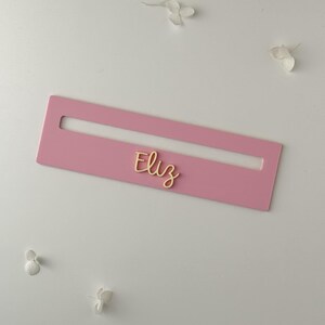 May include: A pink rectangular nameplate with a cutout, featuring the name "Eliz" in gold script. The nameplate is set against a white background with scattered white floral accents.