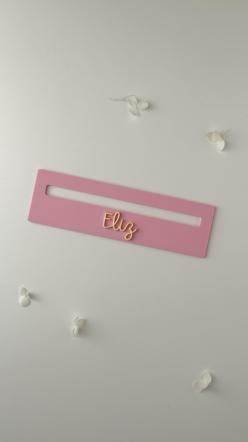 May include: A pink rectangular nameplate with a cutout, featuring the name "Eliz" in gold script. The nameplate is set against a white background with scattered white floral accents.