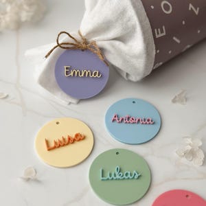 Könnte beinhalten: Eine Sammlung farbenfroher, runder Namensschilder mit Namen in Schreibschrift. Die Schilder sind in verschiedenen Pastellfarben gehalten, darunter Lavendel, Gelb, Blau und Grün. Ein Schild ist an einem weißen Stoffbündel befestigt, das mit Bindfaden zusammengebunden ist.