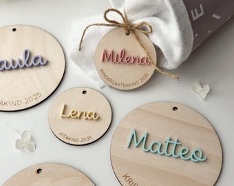 May include: A collection of wooden name tags with 3D lettering in various colours. The tags, featuring names such as "Milena" and "Matteo", are round and accompanied by additional text. A drawstring bag is in the background.