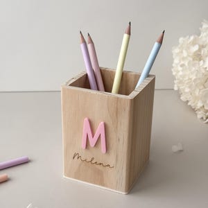 Könnte beinhalten: Ein Bleistifthalter aus Holz mit dem Buchstaben "M" und dem Namen "Milena" auf der Vorderseite. Der Halter ist mit vier Bleistiften in Pastellfarben gefüllt: hellblau, rosa, lavendel und gelb.