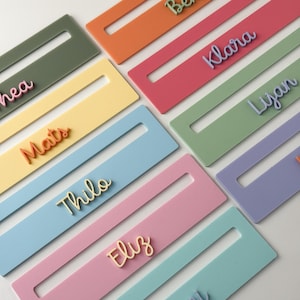 May include: A selection of pastel-coloured name tags, each with a different name in a contrasting hue. The tags are rectangular with a central cutout. Colours include pink, blue, and green. The names are in a cursive font, creating a visually appealing arrangement.