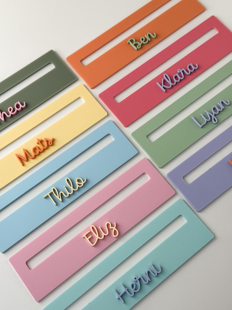 May include: A selection of pastel-coloured name tags, each with a different name in a contrasting hue. The tags are rectangular with a central cutout. Colours include pink, blue, and green. The names are in a cursive font, creating a visually appealing arrangement.