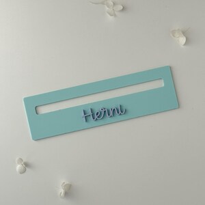 May include: A pale blue rectangular name tag with a cut-out section, displaying the name "Herni" in a cursive script. The tag is set against a white backdrop, with scattered white flower petals.
