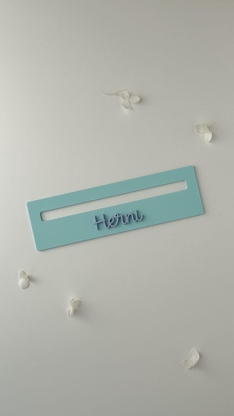 May include: A pale blue rectangular name tag with a cut-out section, displaying the name "Herni" in a cursive script. The tag is set against a white backdrop, with scattered white flower petals.