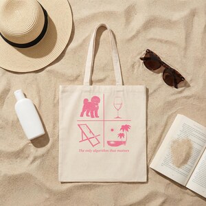 Può includere: Una borsa tote in tela beige con una stampa grafica rosa divisa in quattro quadranti contenente un barboncino, un calice di vino, una sedia a sdraio e palme, con il testo "The only algorithm that matters" stampato sotto il disegno.