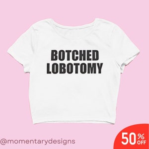 Puede incluir: Top corto blanco con la frase "BOTCHED LOBOTOMY" impresa en letras negras en negrita. La camiseta tiene mangas cortas y cuello redondo. El fondo es de color rosa claro. Una etiqueta de venta roja en la esquina inferior derecha dice "50% OFF".