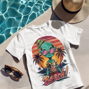 Puede incluir: Camiseta blanca con un alienígena de dibujos animados con gafas de sol y un sombrero, con un diseño de puesta de sol y palmeras. La palabra "Beach!" está escrita en una fuente colorida. También se ven gafas de sol y un sombrero de paja.