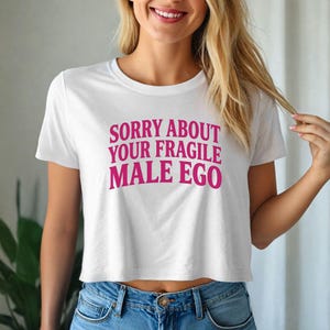 May include: White crop top with the text "SORRY ABOUT YOUR FRAGILE MALE EGO" in pink. The shirt has a crew neck and short sleeves. The model is wearing blue jeans.