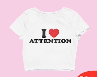 Op de afbeelding: Witte crop top met de tekst "I ♥ ATTENTION" in zwart en rood. De top heeft een ronde hals en korte mouwen. Een rode banner in de rechter benedenhoek toont "50% OFF".