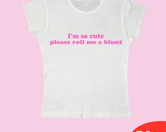 Puede incluir: Camiseta blanca con cuello redondo y mangas cortas. La camiseta tiene el texto rosa "I'm so cute please roll me a blunt". El fondo es de color rosa claro.