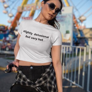 Puede incluir: Un top blanco corto con el texto "slightly delusional but very hot" impreso en la parte delantera. La modelo lleva pantalones cortos negros y una camisa de cuadros atada a la cintura.