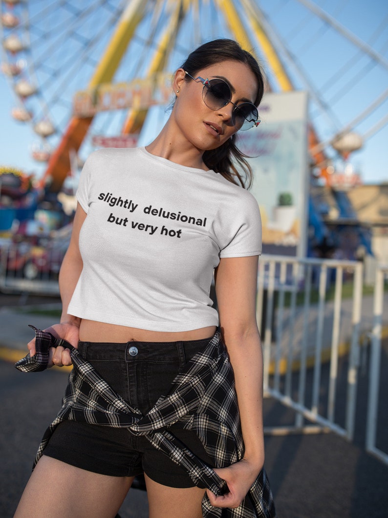 Puede incluir: Un top blanco corto con el texto "slightly delusional but very hot" impreso en la parte delantera. La modelo lleva pantalones cortos negros y una camisa de cuadros atada a la cintura.