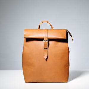 May include: A brown leather backpack with a flap closure and a silver buckle. The backpack has a simple design and is made of high-quality leather.