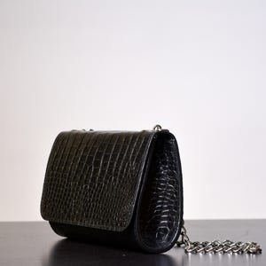 May include: A black, textured handbag with a silver chain strap. The bag features a flap closure and a crocodile-like pattern. The bag is resting on a dark surface.