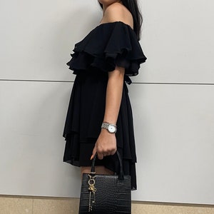 May include: A woman wearing a black off-the-shoulder dress with ruffles and a black crocodile-embossed handbag with a gold key charm.