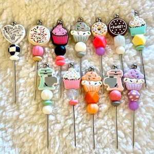 May include: A set of 12 colorful cupcake and baking themed drink stirrers. The stirrers are made of metal with various colorful silicone beads and charms. The charms include cupcakes, a mixer, a donut, and a cookie.