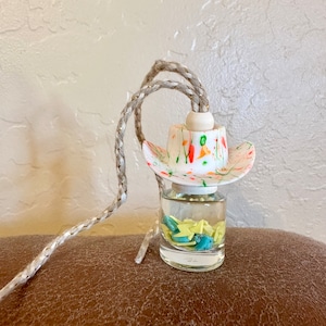 May include: A white cowboy hat with colorful paint splatter sits atop a clear glass bottle filled with turquoise and yellow star-shaped beads. The bottle is attached to a long, beige rope.